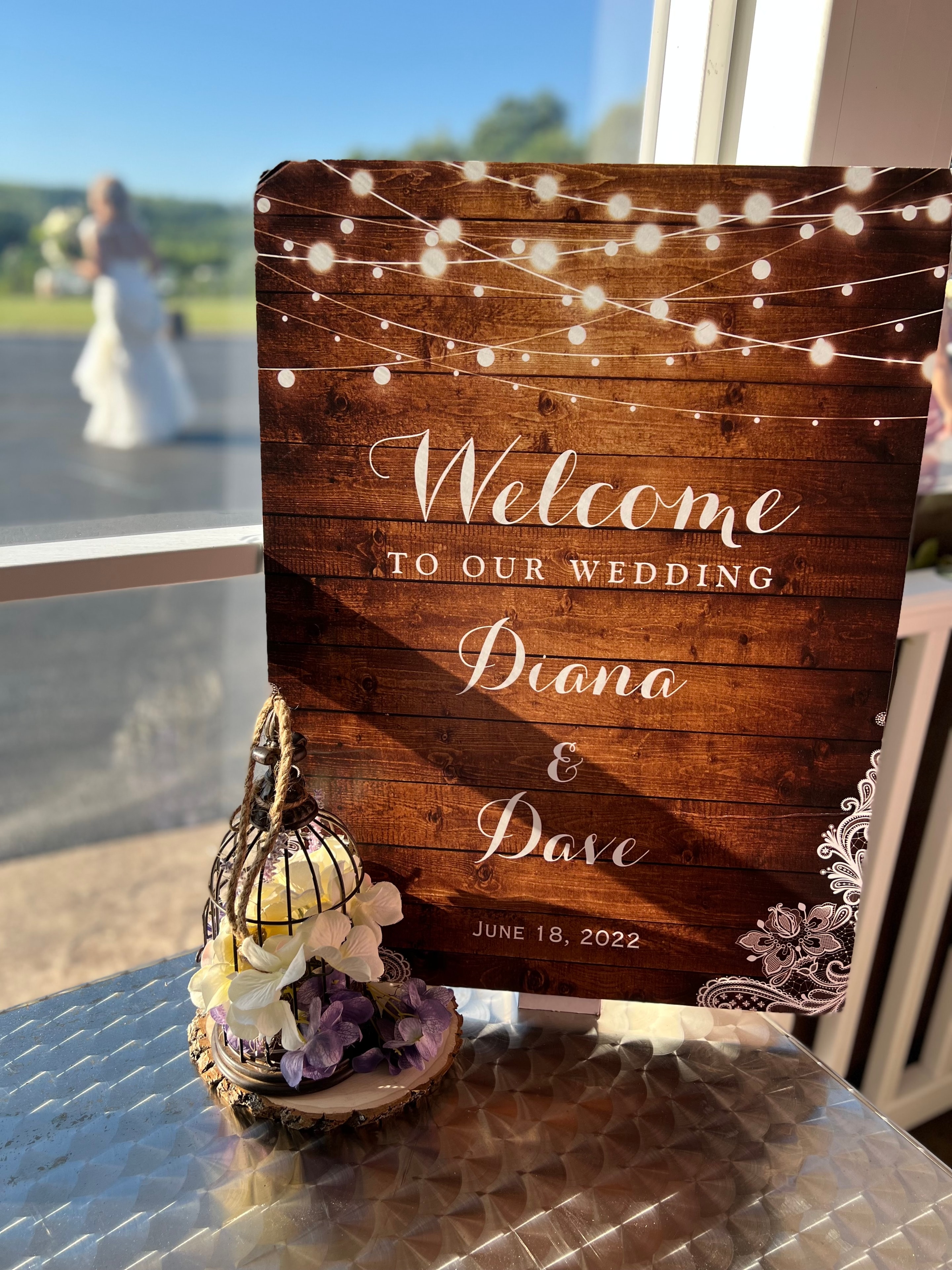 Rustic wooden wedding welcome sign with string lights and bride visible in background